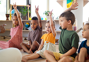 Group of small nursery school children sitting on floor indoors in classroom, raising hands.